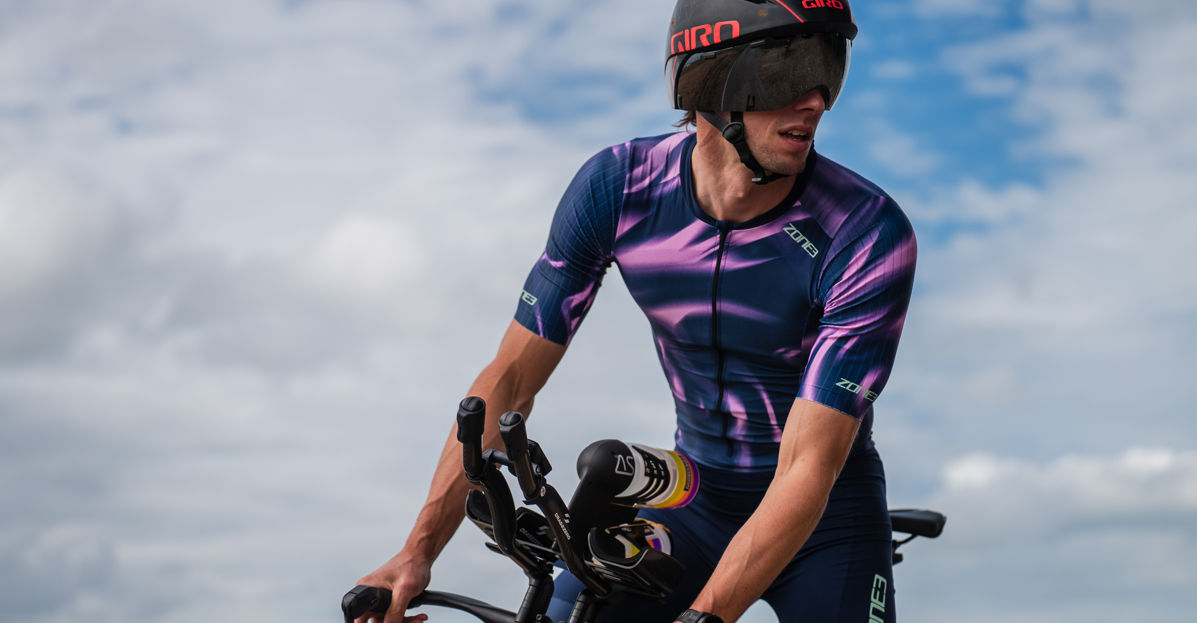 Cyclist in a pink and purple zone3 trisuit riding a bicycle on a road with clouds in the background