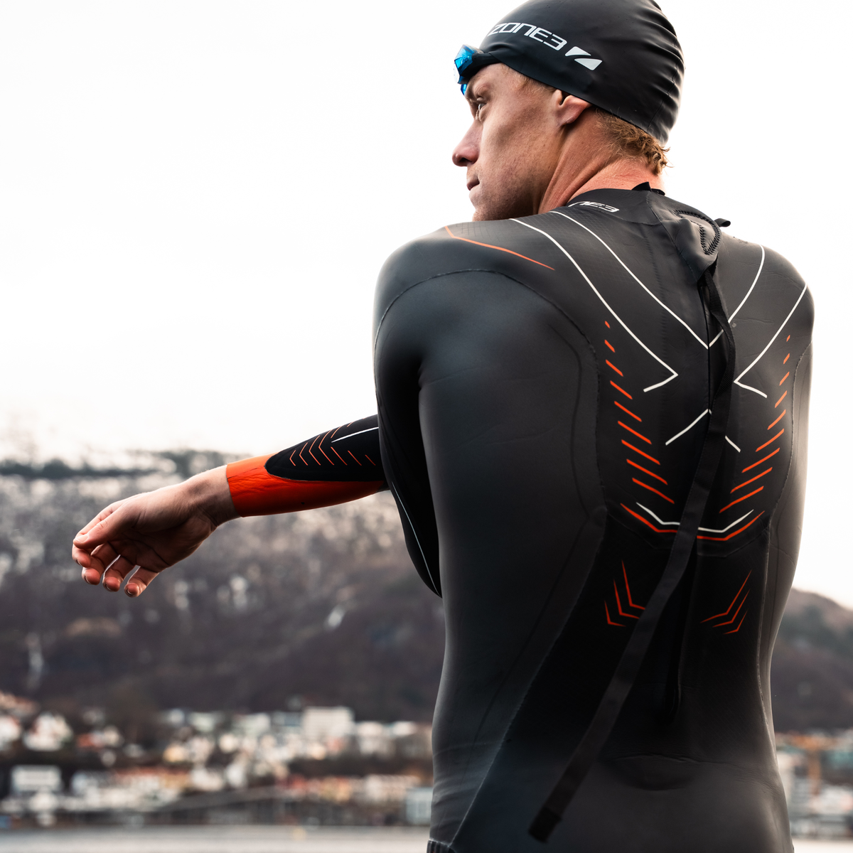 Casper Stornes wearing a black vanquish x wetsuit with red accents, standing outdoors with mountains in the background.