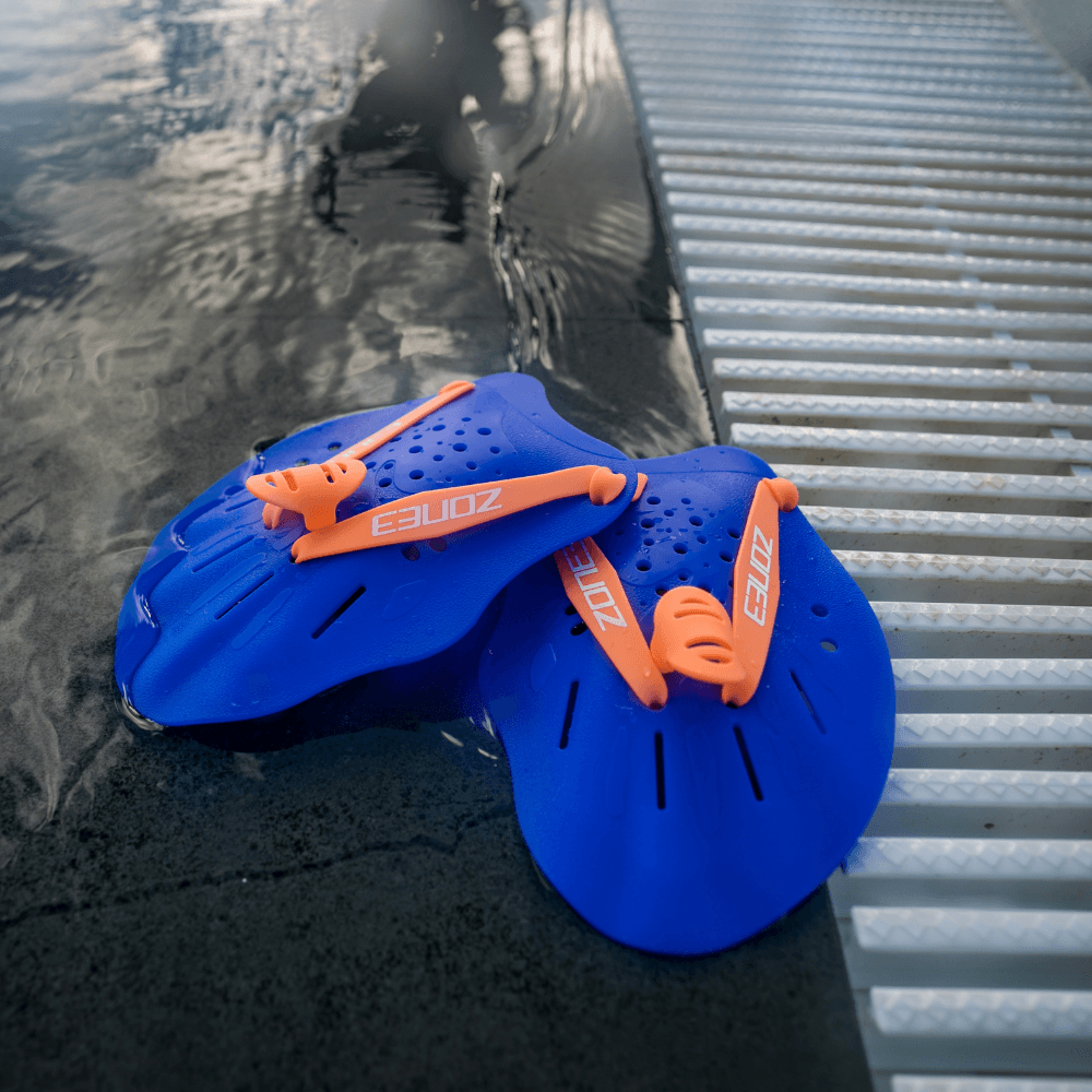 Par de palas de entrenamiento Ergo en azul con correas naranjas, apoyadas en el borde de la piscina junto a una rejilla metálica.