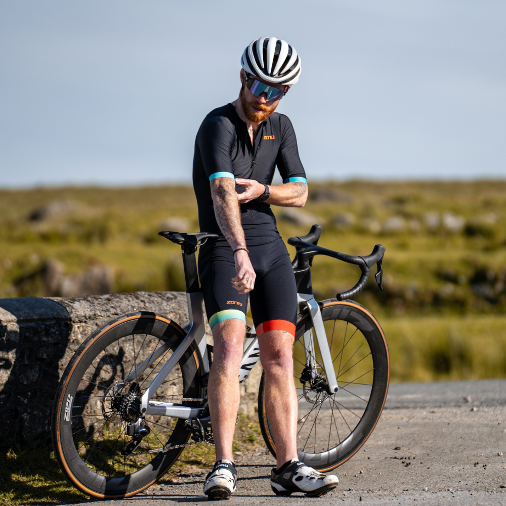 Hombre con el trisuit Activate+ de ZONE3 en negro, de pie junto a una bici de carretera, ajustándose la manga con casco y gafas de sol.