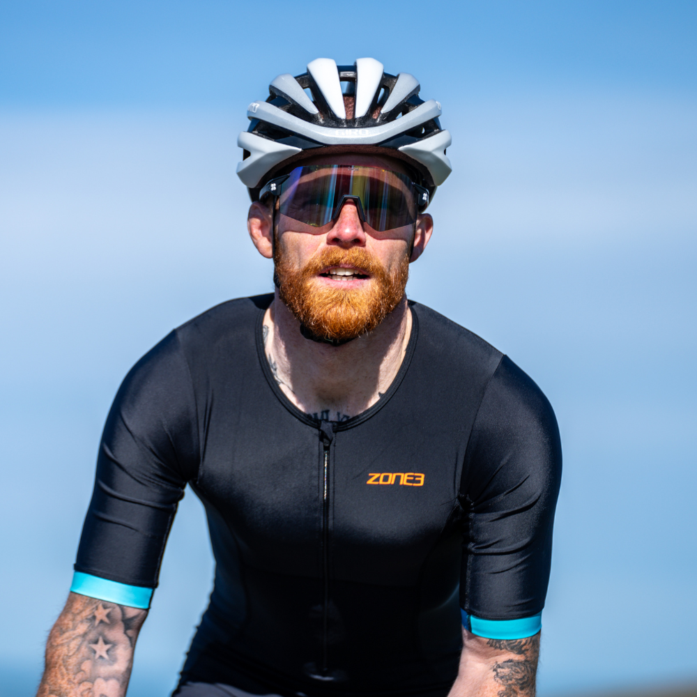 Hombre pedaleando al aire libre con el trisuit Activate+ de manga corta para hombre de ZONE3 en negro, puños de manga azules y logotipo naranja en el pecho.