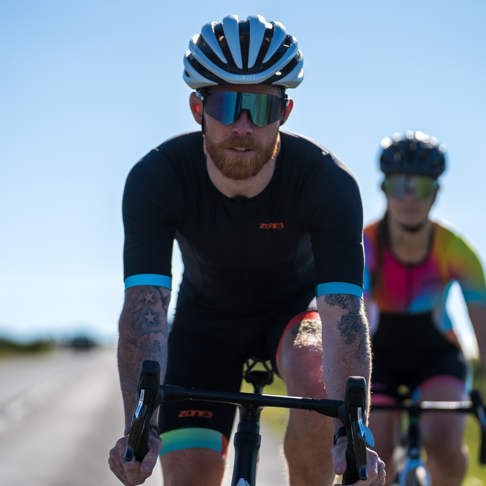 Hombre en bici de carretera con el trisuit Activate+ para hombre de ZONE3 en negro, casco blanco y gafas de sol reflectantes.
