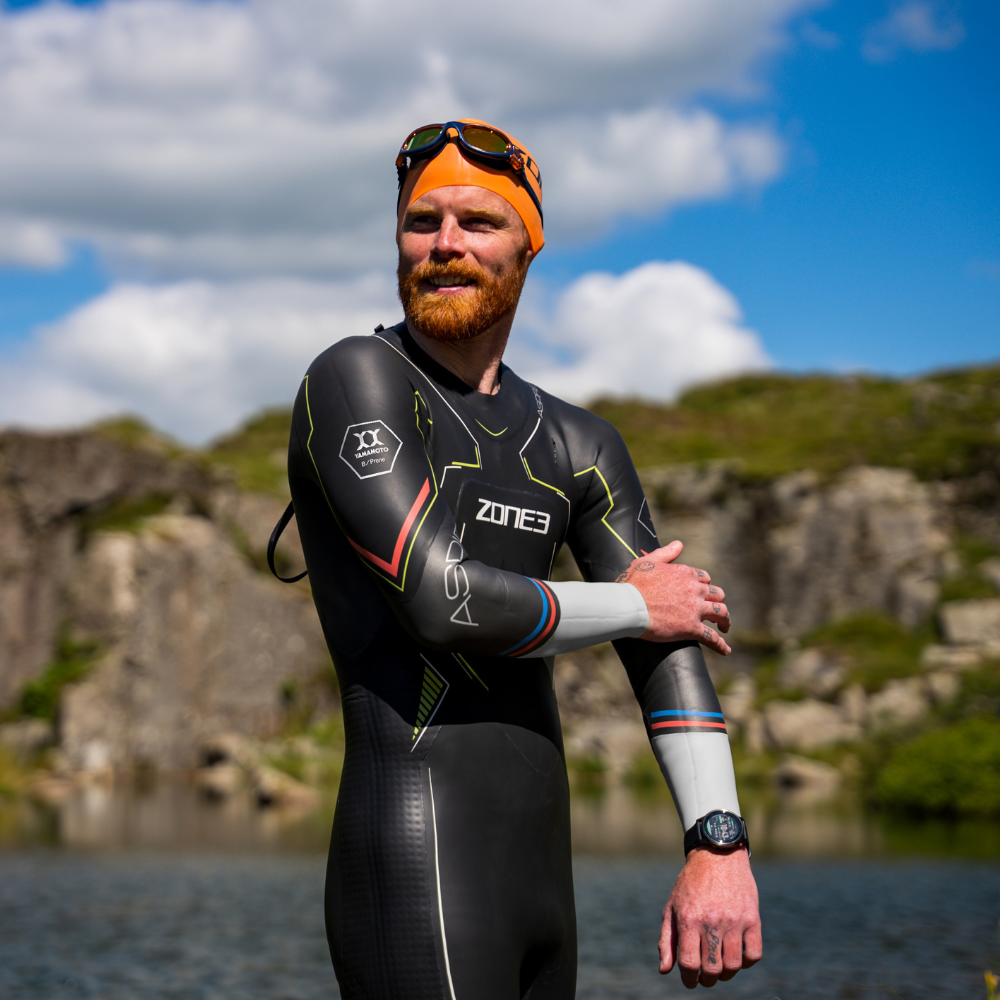 Man wearing a ZONE3 Aspire wetsuit. He has his hand on his left arm and is looking past the camera. There is a body of water behind him and a rocky green backdrop surrounding.