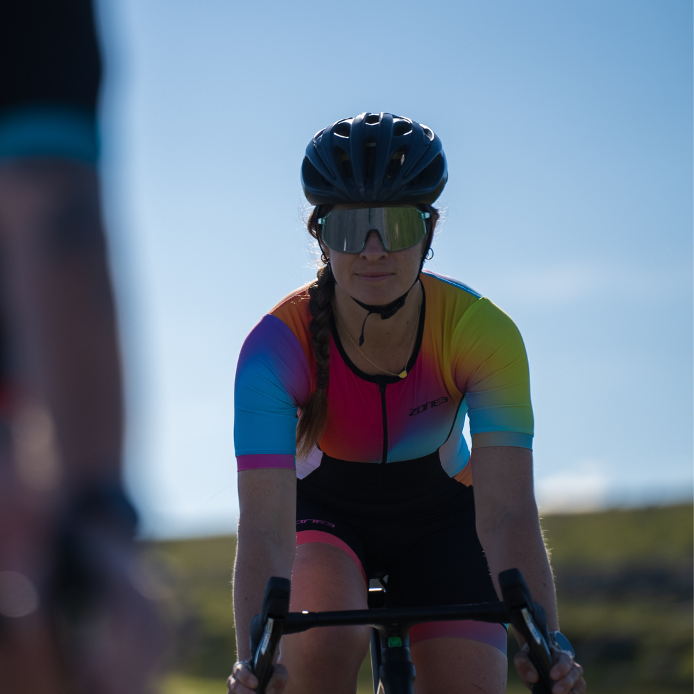 Mujer pedaleando de frente con el trisuit Activate+ de manga corta para mujer de ZONE3, gafas de sol espejadas y casco negro. El diseño degradado se aprecia claramente en el pecho y las mangas.
