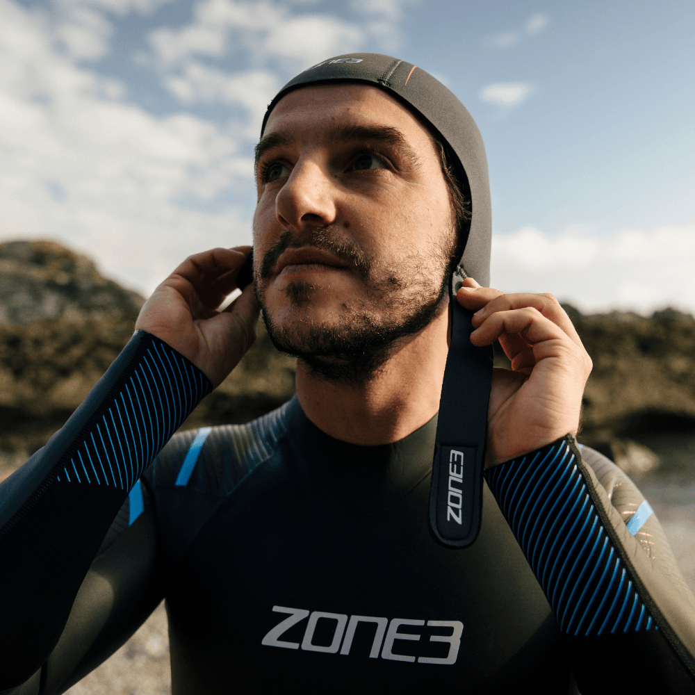 Hombre ajustándose el gorro de natación Active-Flex mientras lleva un neopreno en la playa, perfecto para nadar y hacer surf.