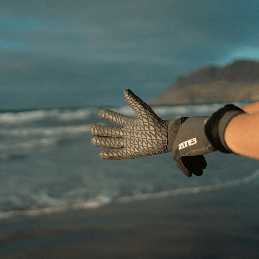 Persona mostrando los guantes de natación Active-Flex en la playa, ideales para nadar y deportes de remo.