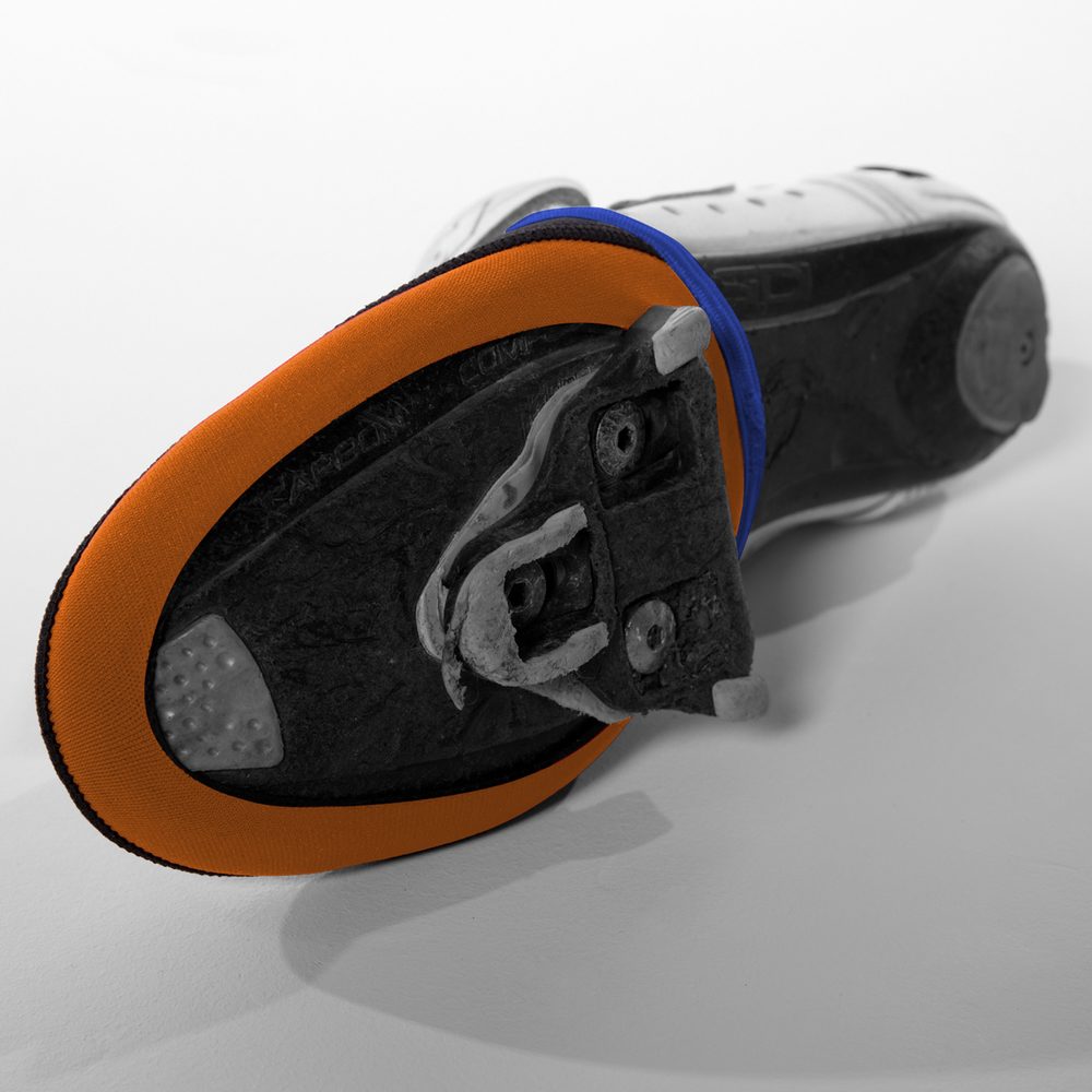 Vista inferior de una zapatilla de ciclismo con el cubrepuntas de neopreno de borde naranja, sujeto a la suela
